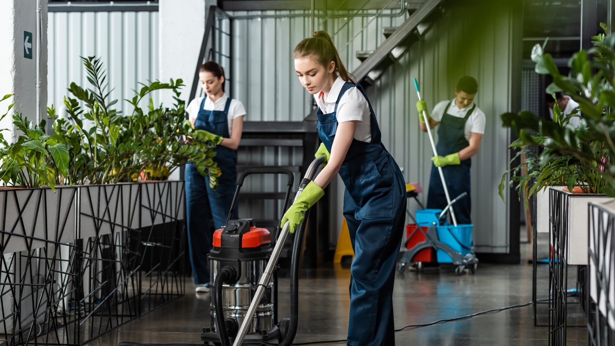 Three individuals in work uniforms and gloves are cleaning an indoor space with plants. One person is using a vacuum cleaner, while the others are engaged in cleaning tasks nearby. The setting features a modern interior with greenery and metal accents.